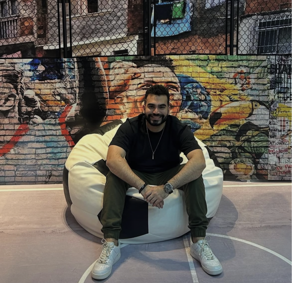 Captura de pantalla 2026-03-04 a la(s) 11.11.57 p. m. Agricultural development volunteer smiling on a bean bag in front of colorful mural.